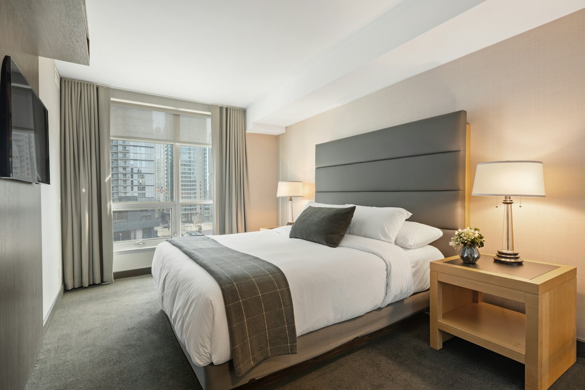 Habitación con cama, mesa de noche de madera y ventanal con vista a la ciudad